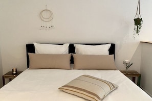 Ein Bett mit weißer Bettwäsche und zwei beigen Kissen, ein Traumfänger an der Wand und eine hängende Pflanze.