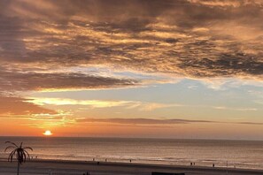 Eine Szene vom Strand bei Sonnenuntergang über dem Meer, eine Palme und einige Personen, die am Ufer entlanggehen.