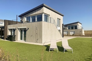 Een modern gebouw met twee verdiepingen, een plat dak, grote ramen en een overdekt terras met een bankje.