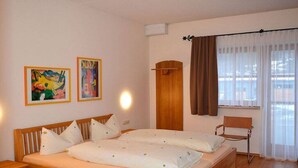Quarto com cama de casal, duas pinturas na parede, guarda-roupa de madeira, uma cadeira e uma janela com cortinas.