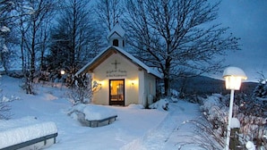 Uma pequena igreja com uma cruz na fachada, rodeada por árvores cobertas de neve e um céu claro.