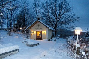 Une petite église surmontée d’une croix, entourée d’arbres couverts de neige, sous un ciel dégagé.