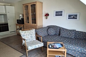 A living room with a sofa, a wooden cabinet, a chair, and a small table with magazines. - Holiday apartment with 1 bedroom (Insel Poel, OT Kirchdorf)