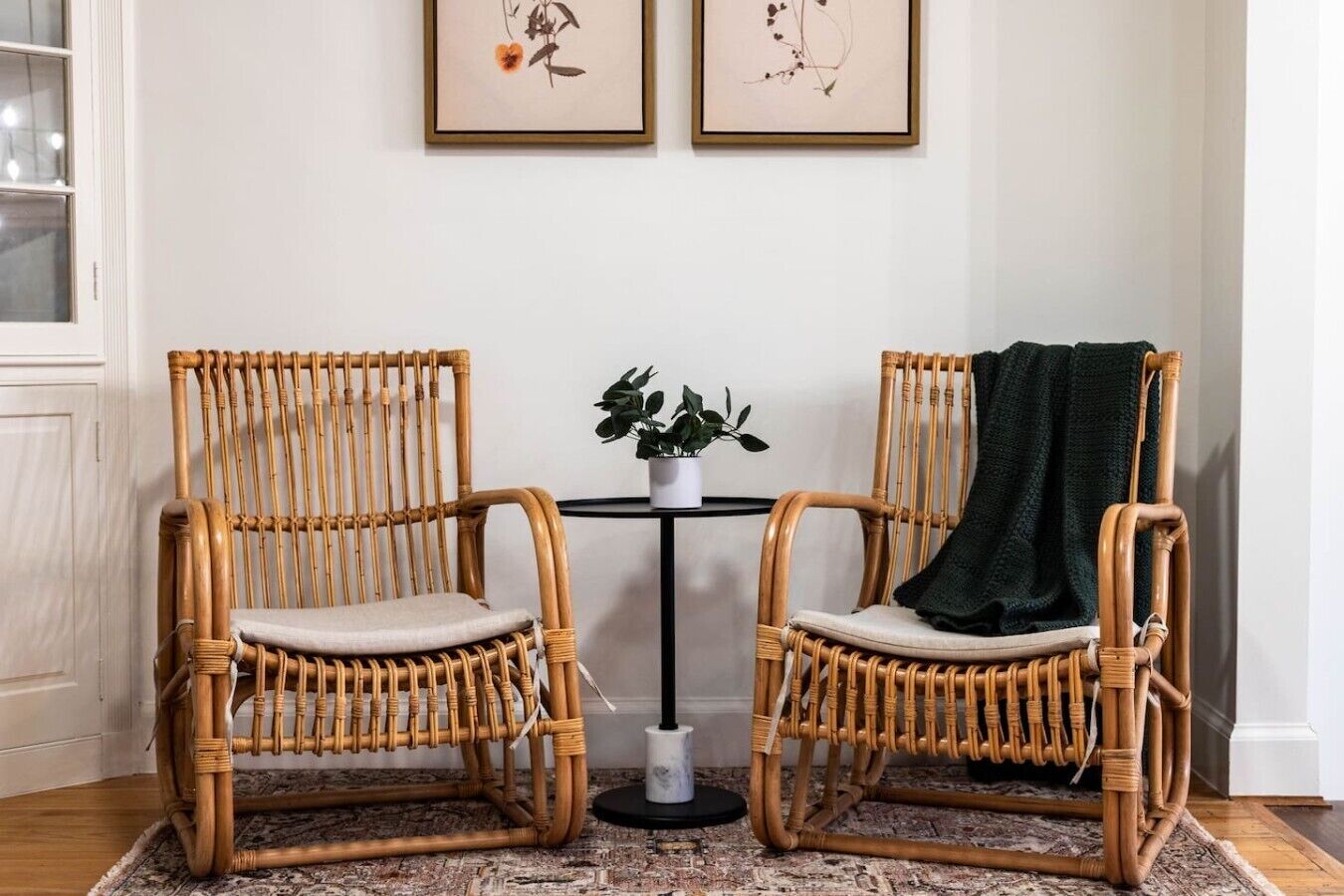 Dos sillas de mimbre con cojines, una mesita redonda con una planta en maceta y un plaid verde oscuro sobre una de las sillas.