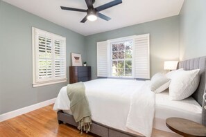 Quarto com cama, ventoinha no teto, duas janelas com persianas, mesa de cabeceira e criado-mudo.