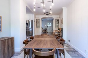 Een ruime eethoek met een lange houten tafel en stoelen, een plafondventilator, en een keuken met roestvrijstalen apparatuur.