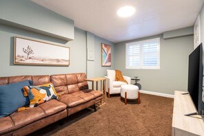 A living room with a brown leather sofa, a white armchair, a small round ottoman, a television on a wooden stand, and artwork on the walls. - Red Butte River Gem-Close to the U, Downtown & Mtn (Salt Lake City)