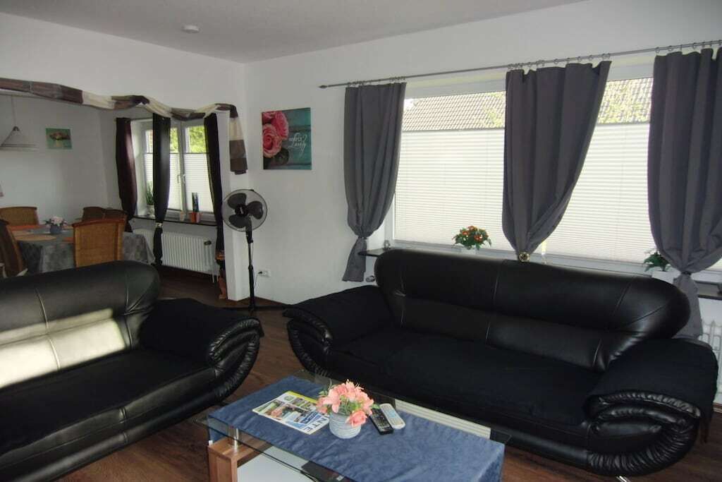 Ein Wohnzimmer mit zwei schwarzen Sofas, einem Couchtisch mit Zeitschriften und einer Vase, einem Standventilator und einem Fenster mit Vorhängen.