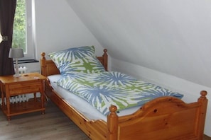 A single wooden bed with a leaf-patterned bedspread in a room with white walls and a window. - Holiday home in Hinte with swimming pool (Hinte)