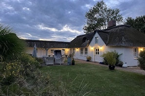 Une maison traditionnelle à toit de chaume, dotée d’une pelouse bien entretenue et d’un espace extérieur aménagé pour s’asseoir.