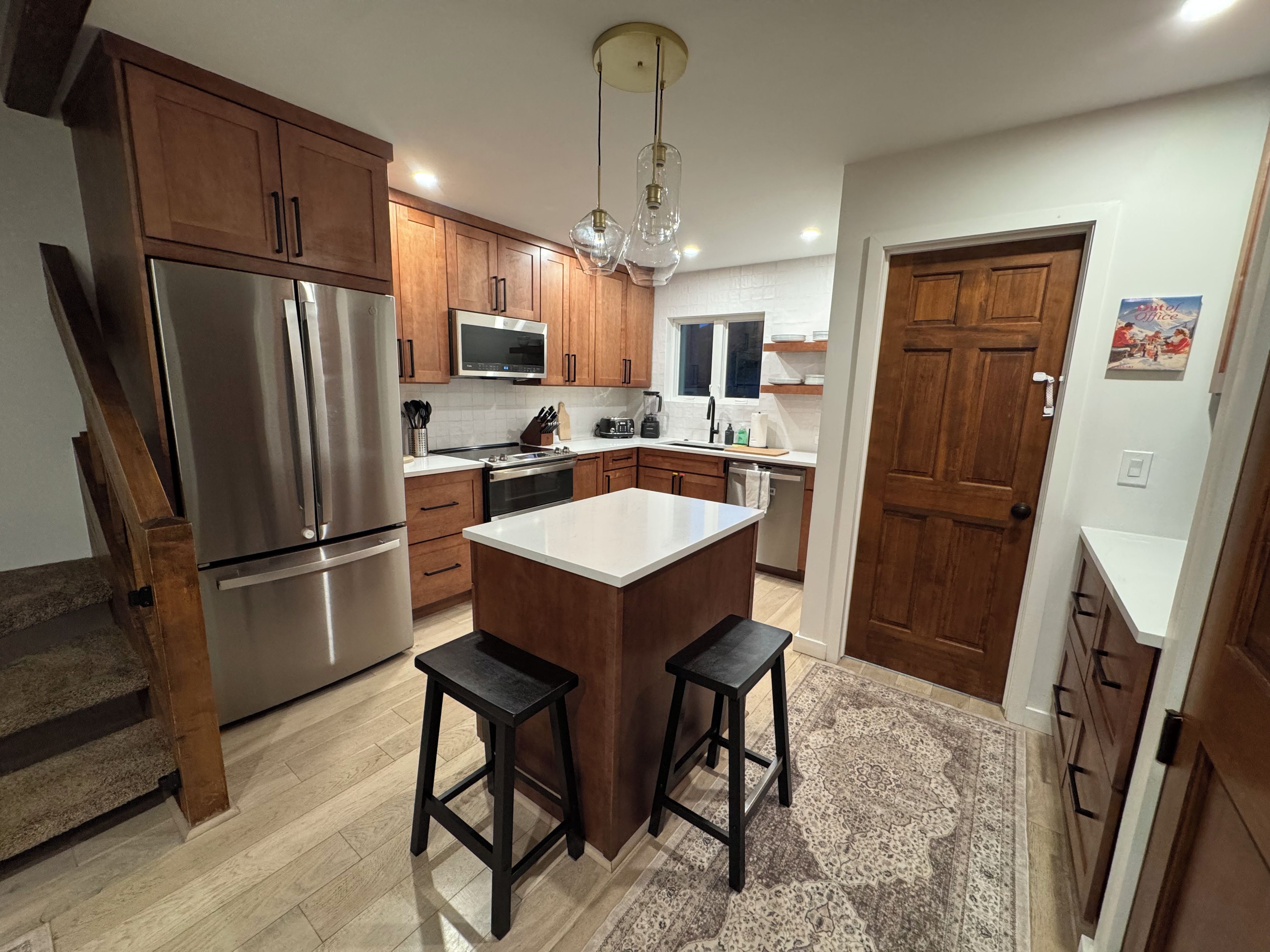 A modern kitchen with wooden cabinets, stainless steel appliances, a central island with stools, and a patterned rug.
