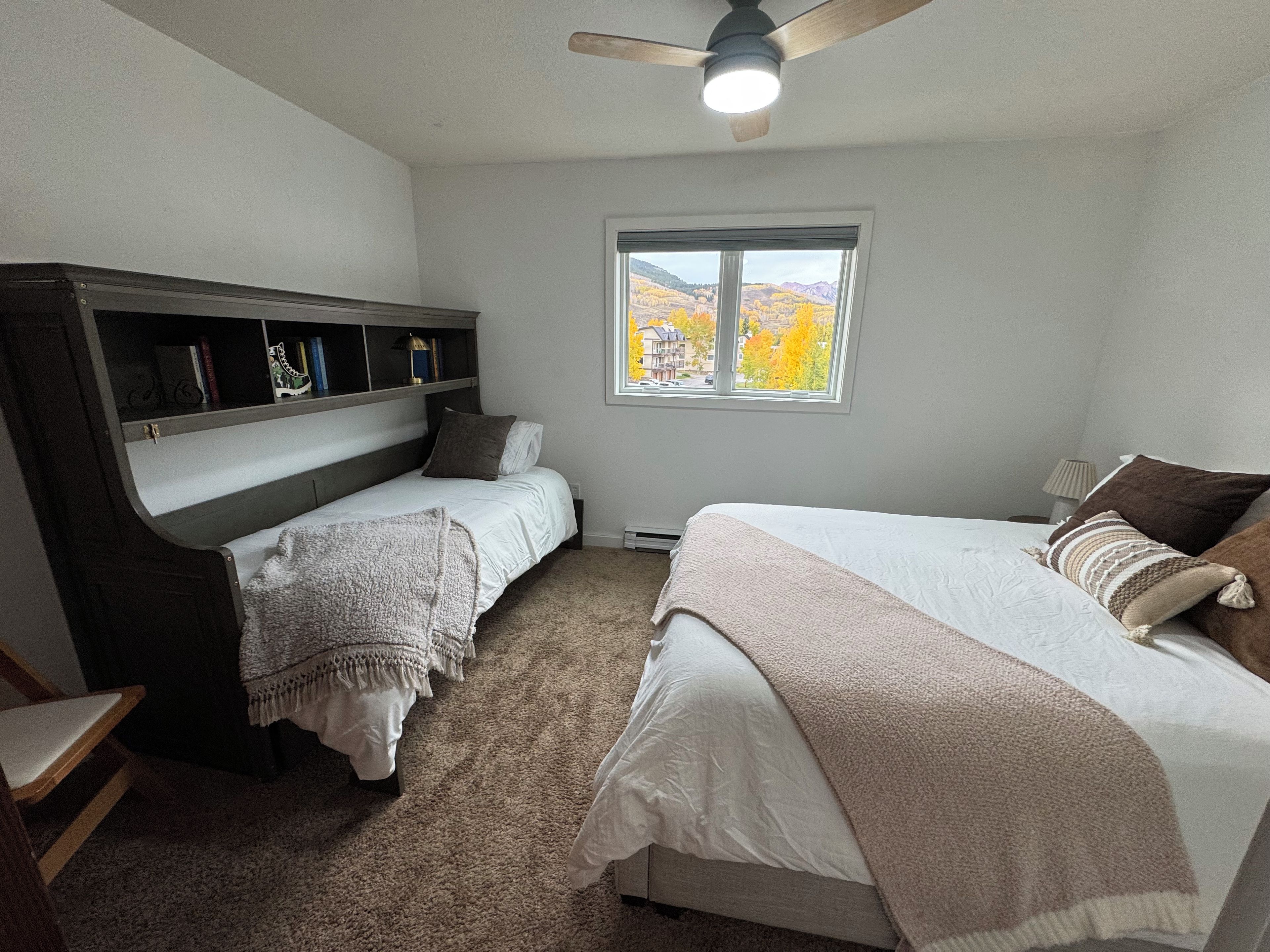 Quarto com duas camas, ventoinha no teto, janela com vista para árvores de outono e uma estante de livros.