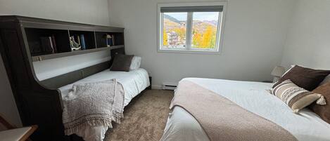 Quarto com duas camas, ventoinha no teto, janela com vista para árvores de outono e uma estante de livros.