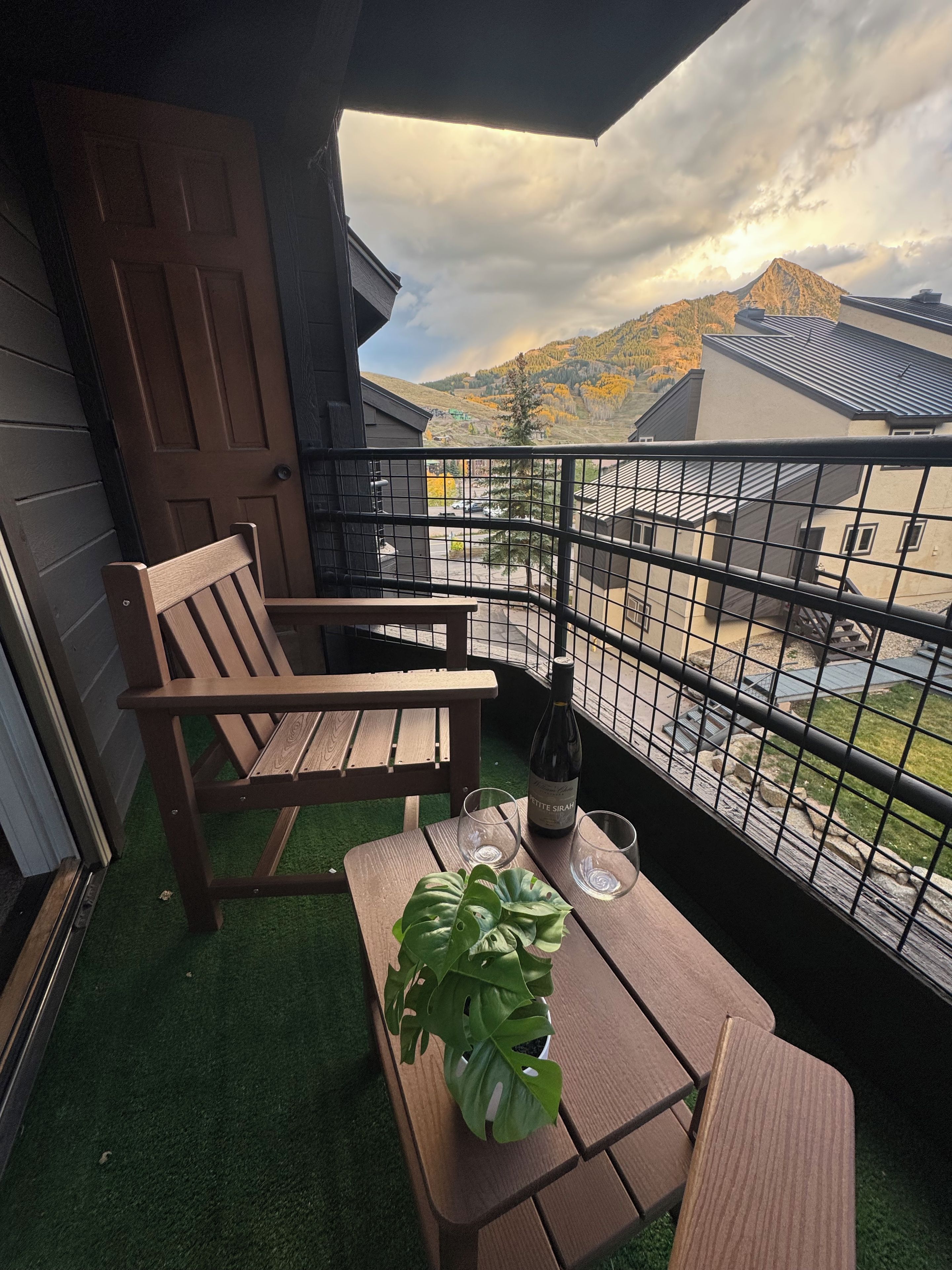 A balcony with a wooden chair, a table with a bottle and glasses, and a view of a mountain and buildings.
