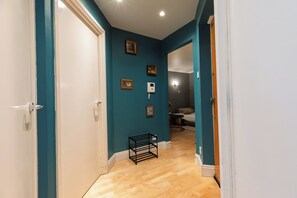 A hallway with teal walls, wooden flooring, and a door leading to a bedroom with a bed and a small table. - Central London 1BR | Trafalgar Square | Sleeps 4 (London)