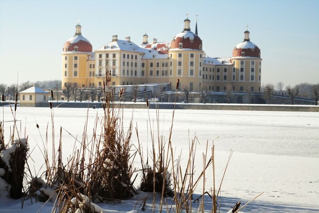 A large, historic building with multiple domes and a symmetrical facade, situated across a frozen body of water.