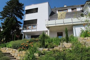 Ein modernes, zweistöckiges Haus mit einem Balkon, umgeben von einem angelegten Garten mit verschiedenen Pflanzen und Blumen.