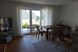Ein Wohnzimmer mit Fernseher, einem Esstisch und Stühlen. Eine Glastür öffnet sich zu einem Außenbereich.
