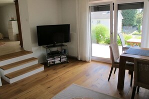 Ein Wohnzimmer mit Flachbildfernseher auf einem Schrank, Holzboden, einem Essbereich mit Tisch und Stühlen sowie einer Glastür zum Garten.