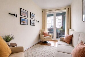 Ein Wohnzimmer mit einer Couch, einem Sessel und einem kleinen Tisch mit einer Pflanze. An der Wand befinden sich gerahmte Bilder und eine Glastür zum Außenbereich.