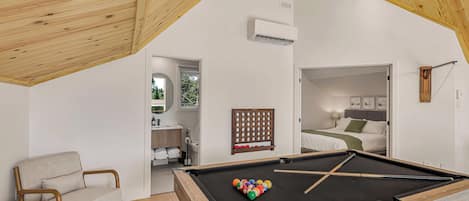 A modern room with a pool table, a chair, a bed, and a bathroom visible through an open door.
