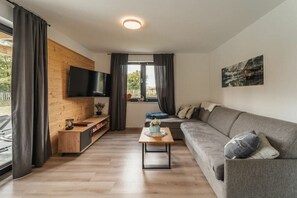 Ein modernes Wohnzimmer mit einer Holzvertäfelung, einem Flachbildfernseher, einem grauen Sofa, einem Couchtisch und einem Fenster mit Vorhängen.