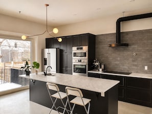 A modern kitchen with a central island, black cabinetry, stainless steel appliances, and a large window.
