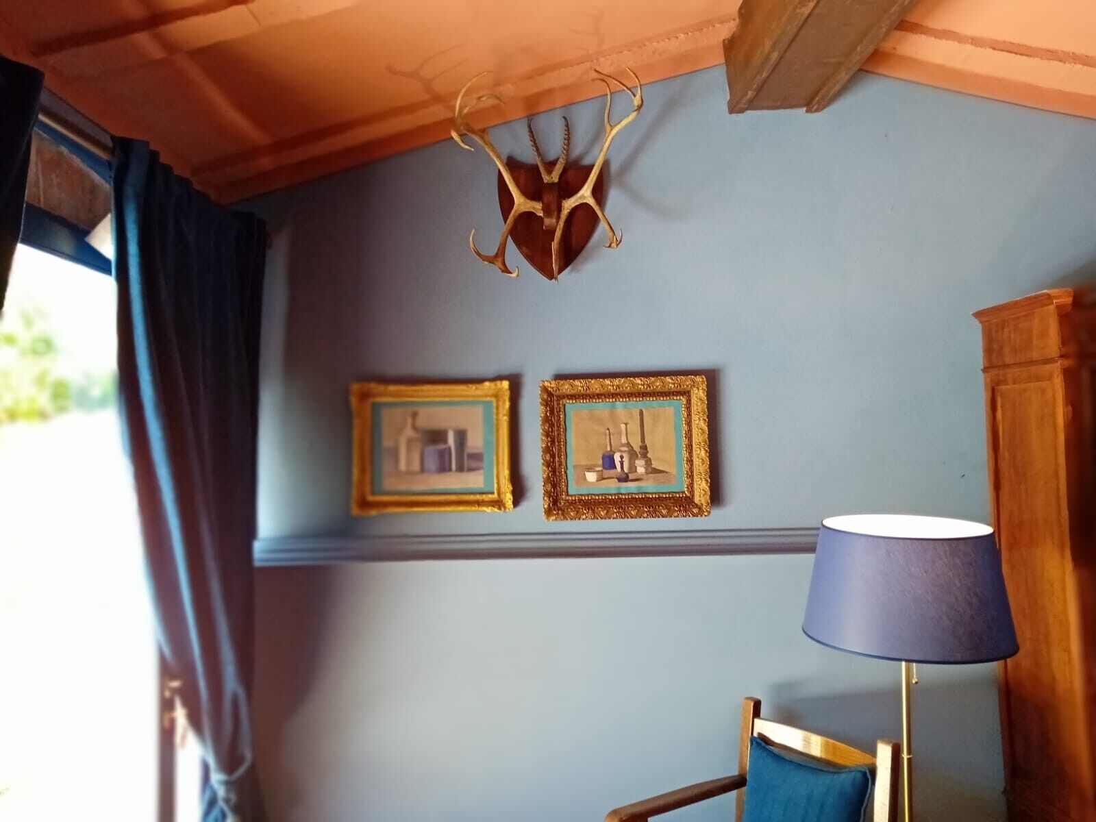 Una habitación con techo de madera, una decoración de cornamenta de ciervo, dos cuadros enmarcados, una lámpara azul y una silla de madera.