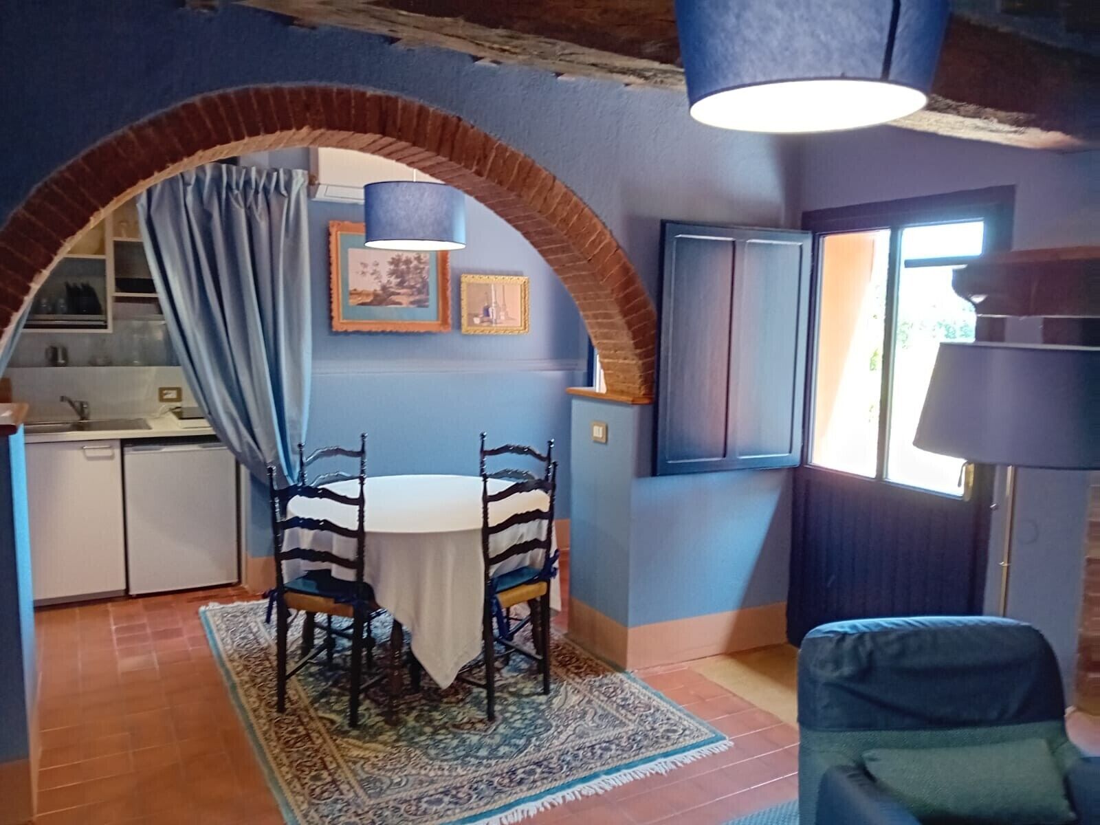 A dining area with a round table and chairs, a patterned rug, a brick archway, and a window with curtains.