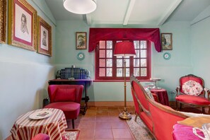 Ein Zimmer mit einem roten Sessel, einem Tisch mit gestreifter Tischdecke, einer Lampe und einem Bücherregal mit Büchern.
