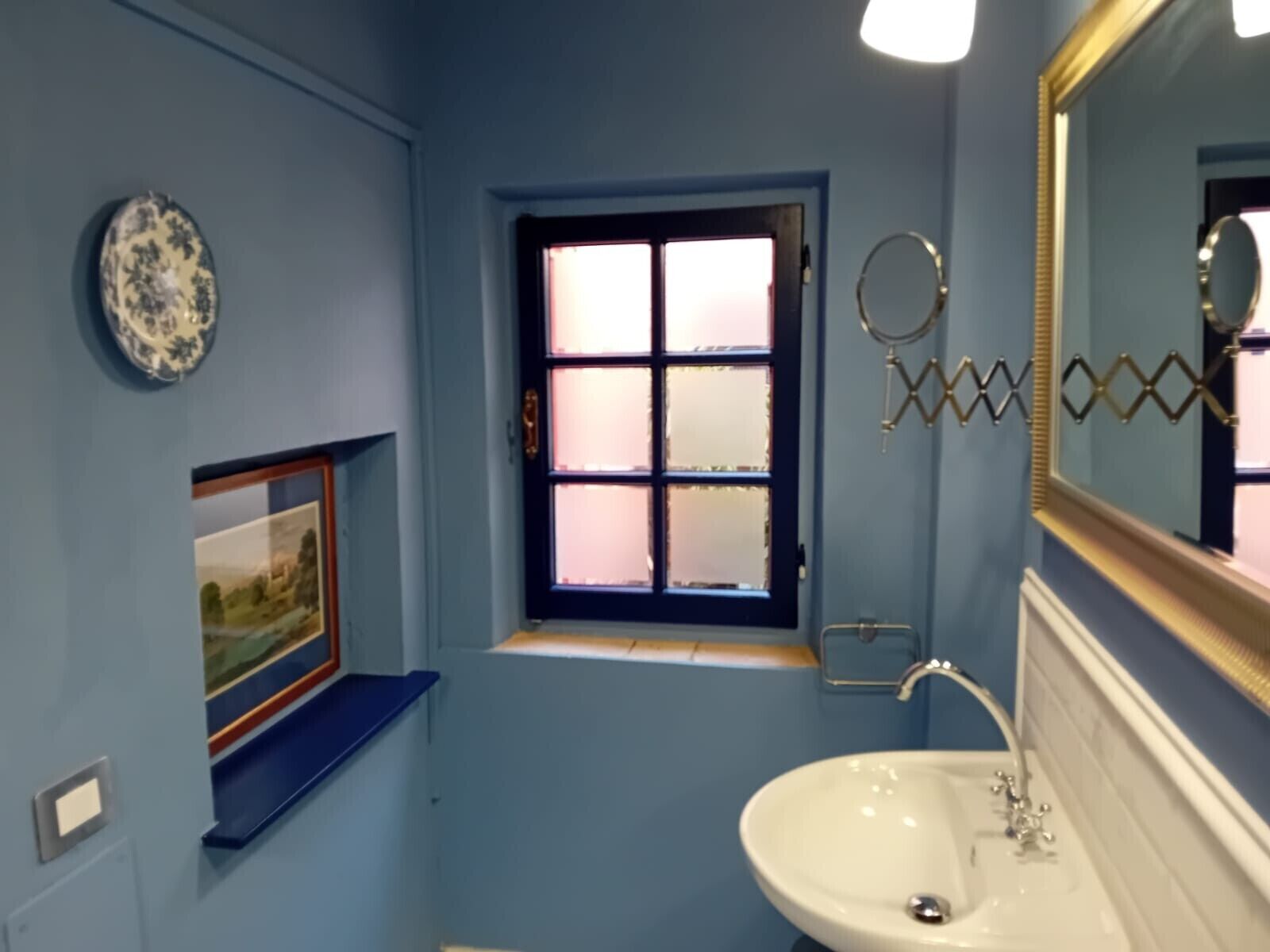 Un baño con pared azul, un lavamanos blanco, un ventanal con vidrio esmerilado, un plato decorativo y un cuadro enmarcado.