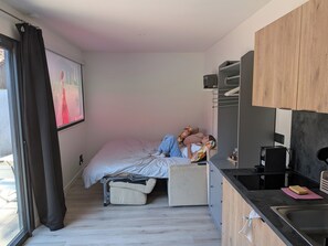 Ein kleines Schlafzimmer mit Bett, einer Sitzecke, einer Küchenzeile und einem Fenster mit Vorhängen.