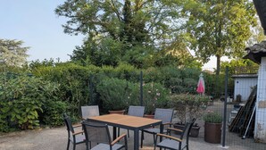 An outdoor patio area with a wooden table and chairs, surrounded by dense greenery and a fence.