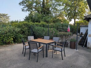 An outdoor patio area with a wooden table and chairs, surrounded by dense greenery and a fence.