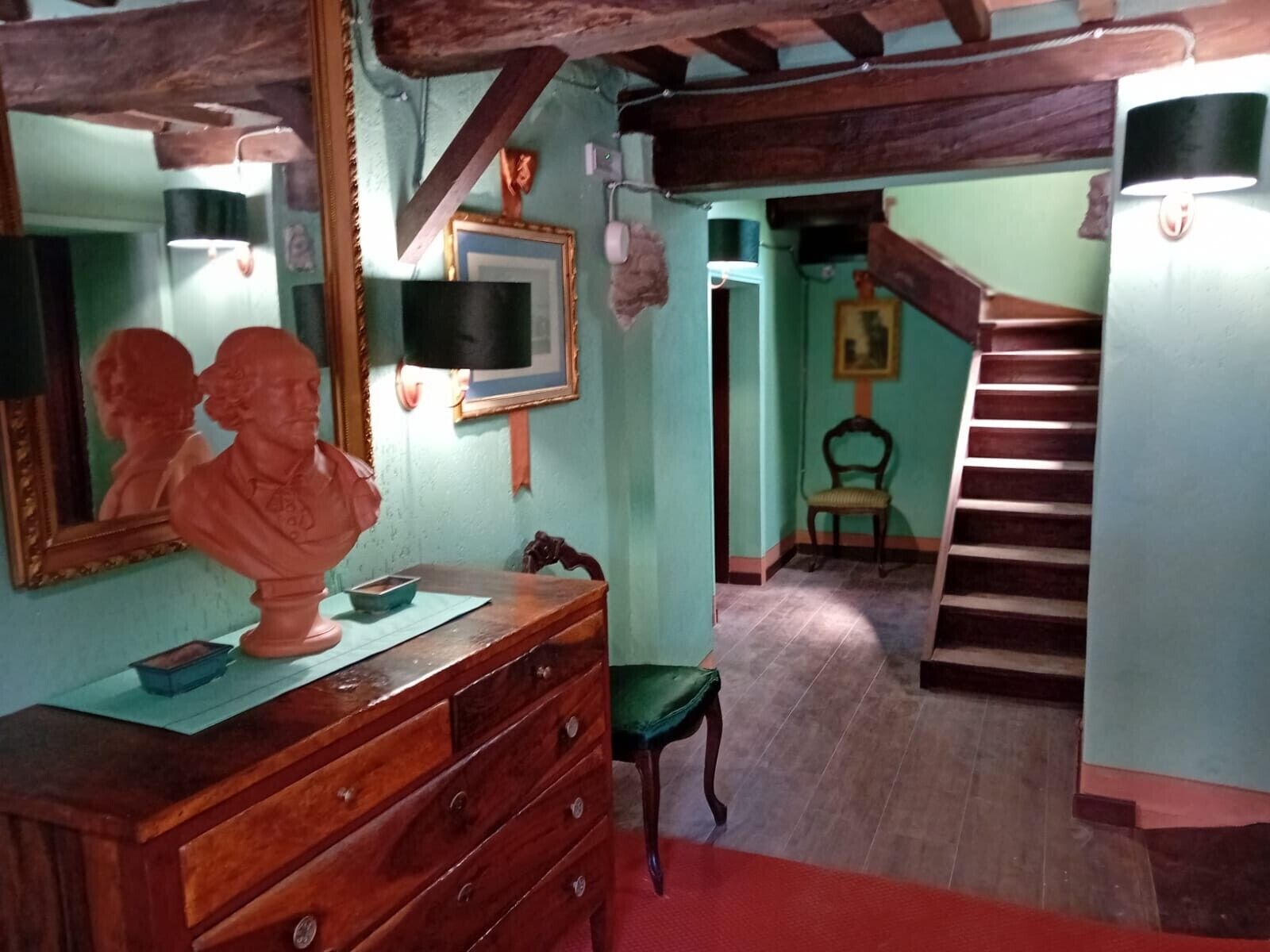 Une pièce comprenant une commode en bois, un buste sculpté, un tableau encadré, une chaise et un escalier.
