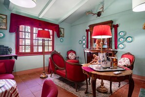 Um quarto com uma poltrona vermelha, uma mesa de madeira com luminária, lareira e uma janela com cortinas.