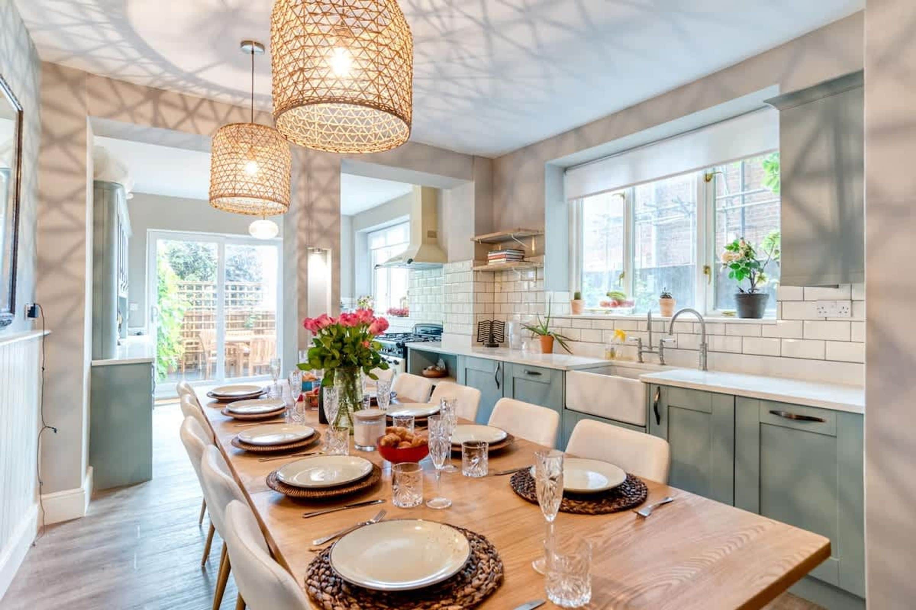 A modern kitchen with a dining area set for a meal, featuring a long table, chairs, and hanging light fixtures.