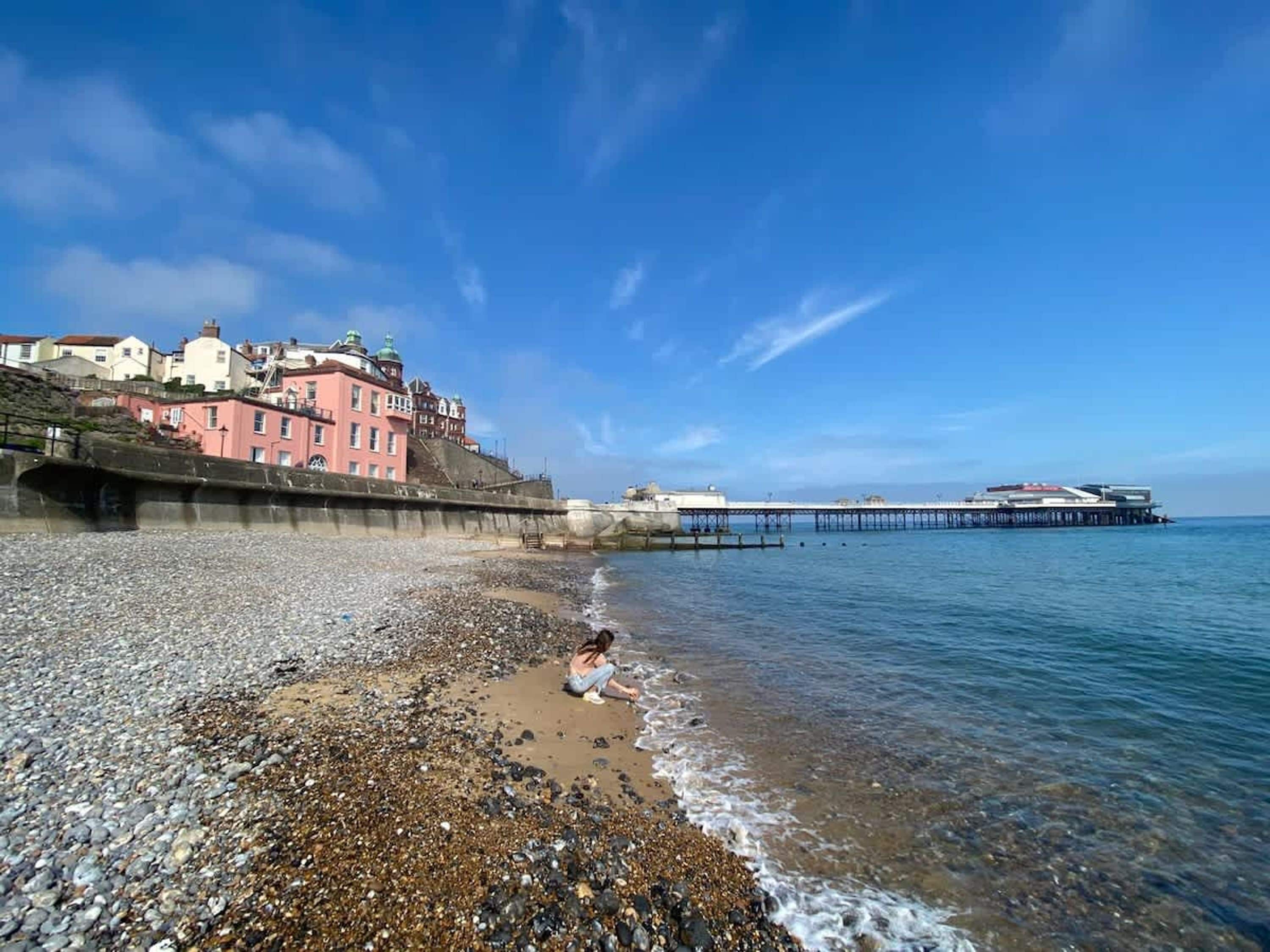 A pebbled beach with a pier extending into the sea, buildings in the background, and a person sitting on the sand.