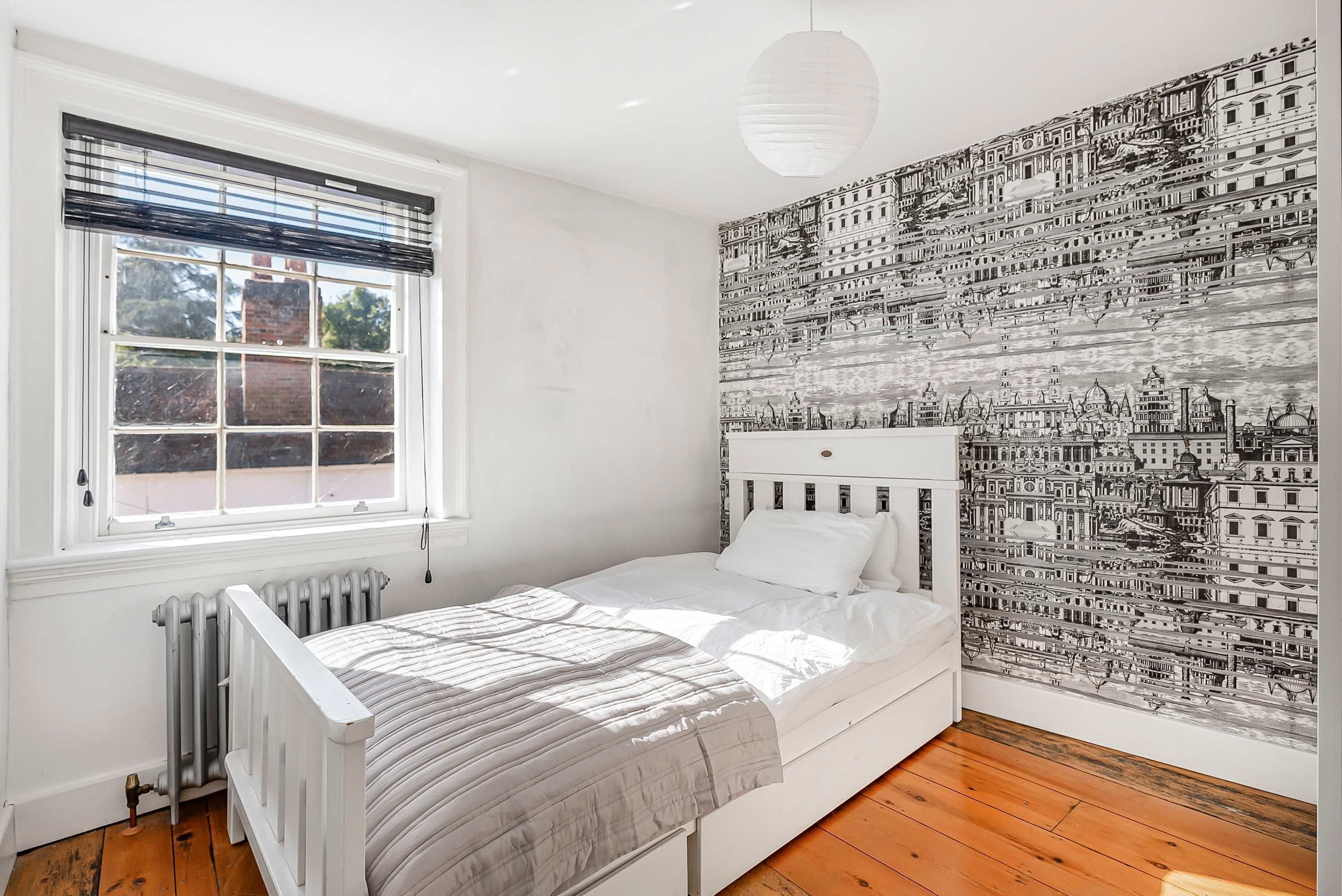 A bedroom with a single bed, a radiator, a window with blinds, and a wall with a detailed cityscape mural.