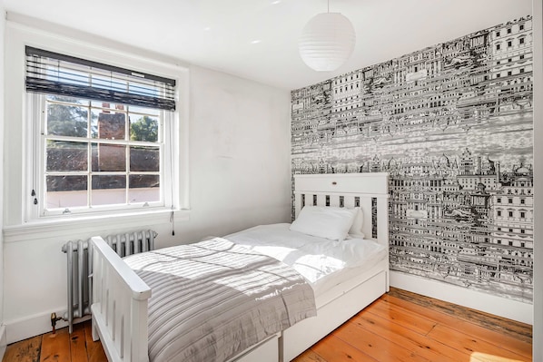 A bedroom with a single bed, a radiator, a window with blinds, and a wall with a detailed cityscape mural.