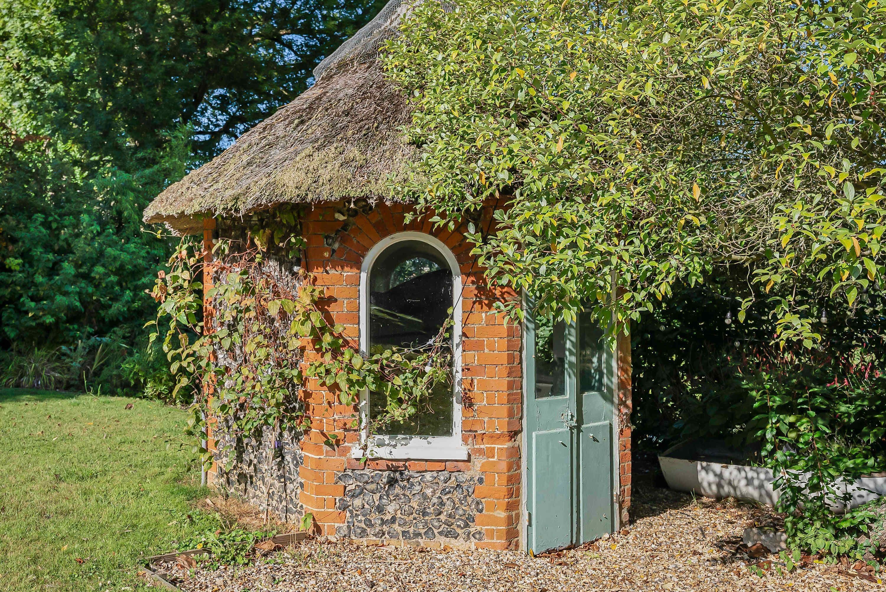 A small brick structure with a thatched roof, a window, and a door, surrounded by greenery.