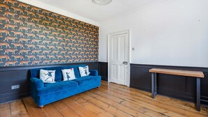Una stanza con un divano blu, carta da parati fantasia, pavimento in legno, una porta bianca e un piccolo tavolino in legno.
