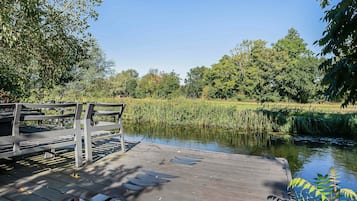 En træbro strækker sig ud i en rolig flod, med frodig vegetation og træer i baggrunden.