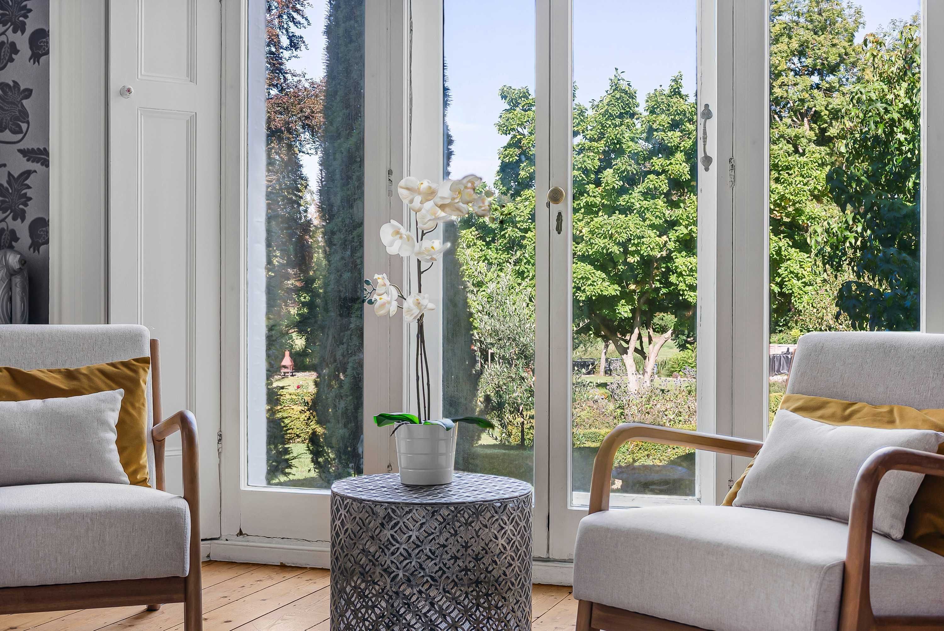A room with two chairs, a small round table, and a view of a garden through large windows.