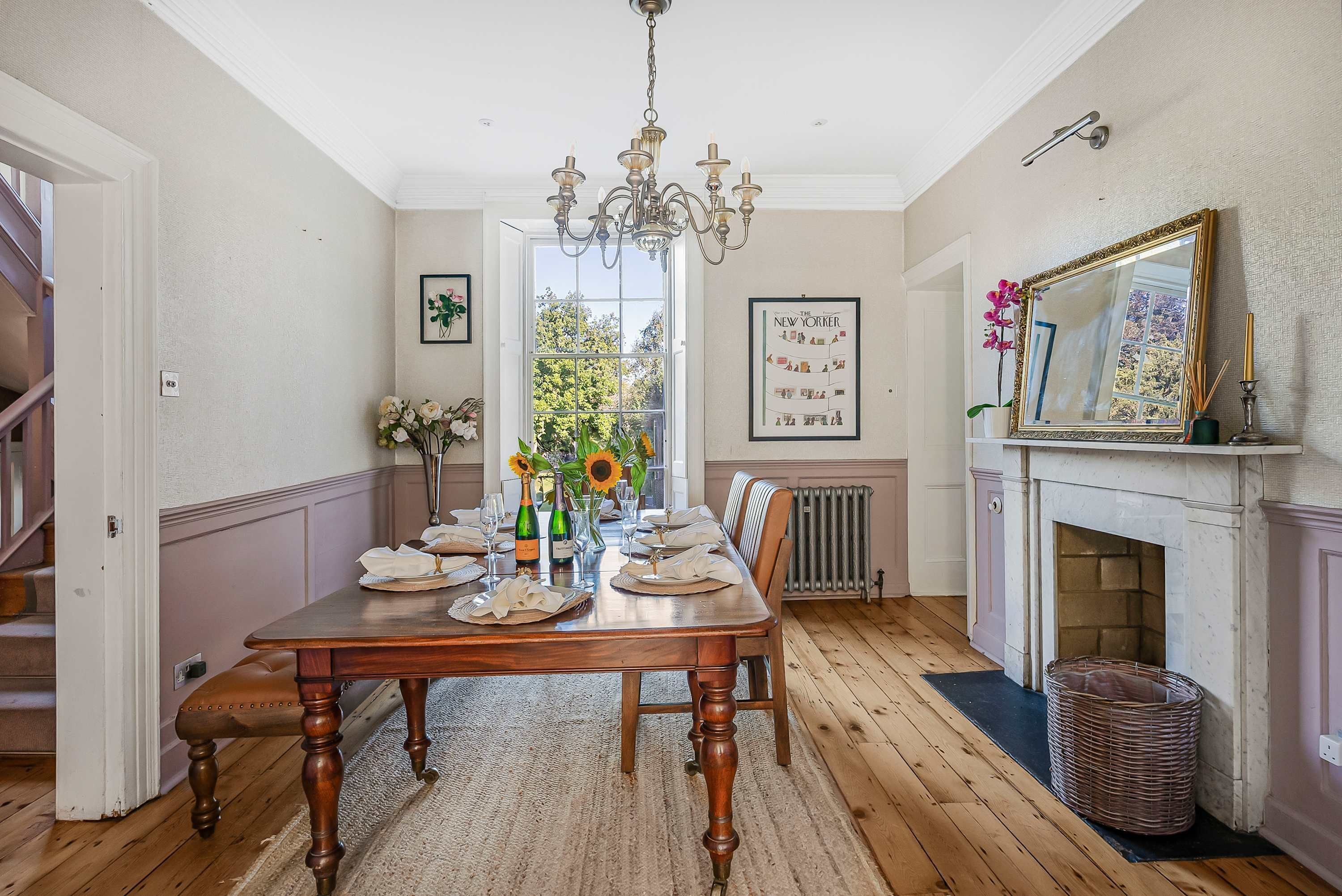 A dining room with a wooden table set for six, a chandelier, a fireplace, and a large window with a view of trees.