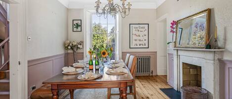 A dining room with a wooden table set for six, a chandelier, a fireplace, and a large window with a view of trees.