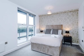 A modern bedroom with a large window, a bed with white linens, a textured wall, and two bedside lamps. - Breathtaking Skyline Views Wi-fi Great Value (Salford)