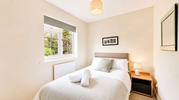 Un dormitorio con cama, ventana, radiador, mesita de noche con lámpara y un cuadro en la pared.