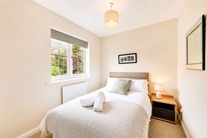 Quarto com cama, janela, radiador, mesa de cabeceira com abajur e quadro na parede.