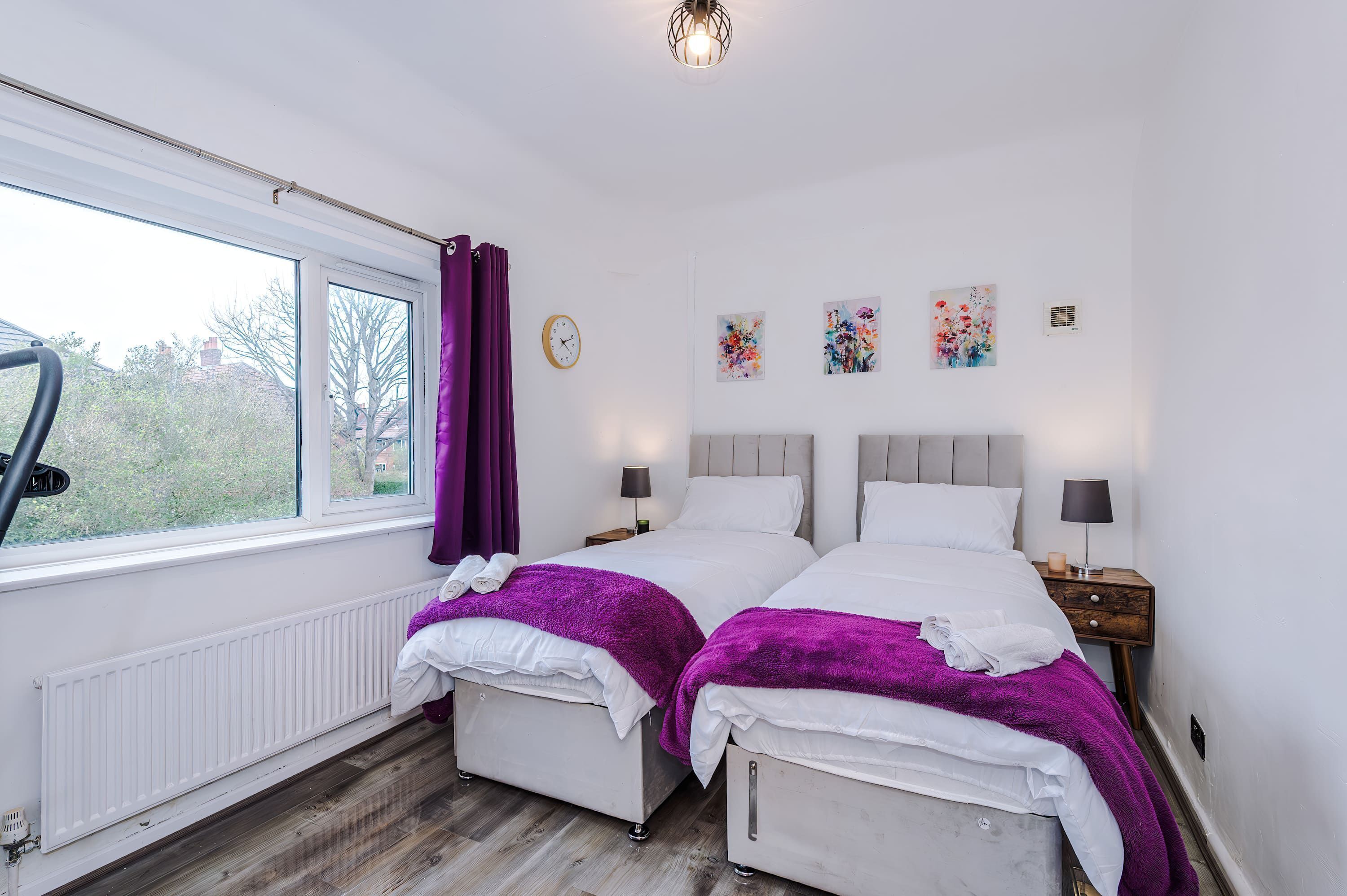 A hotel room with two beds, a window with curtains, a radiator, a clock, and artwork on the wall.